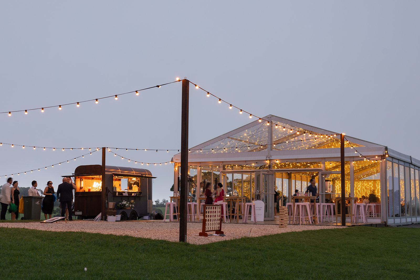 Guests on the gravel at the front of the marquee at dusk, festoon lights overhead and a caravan bar lit up