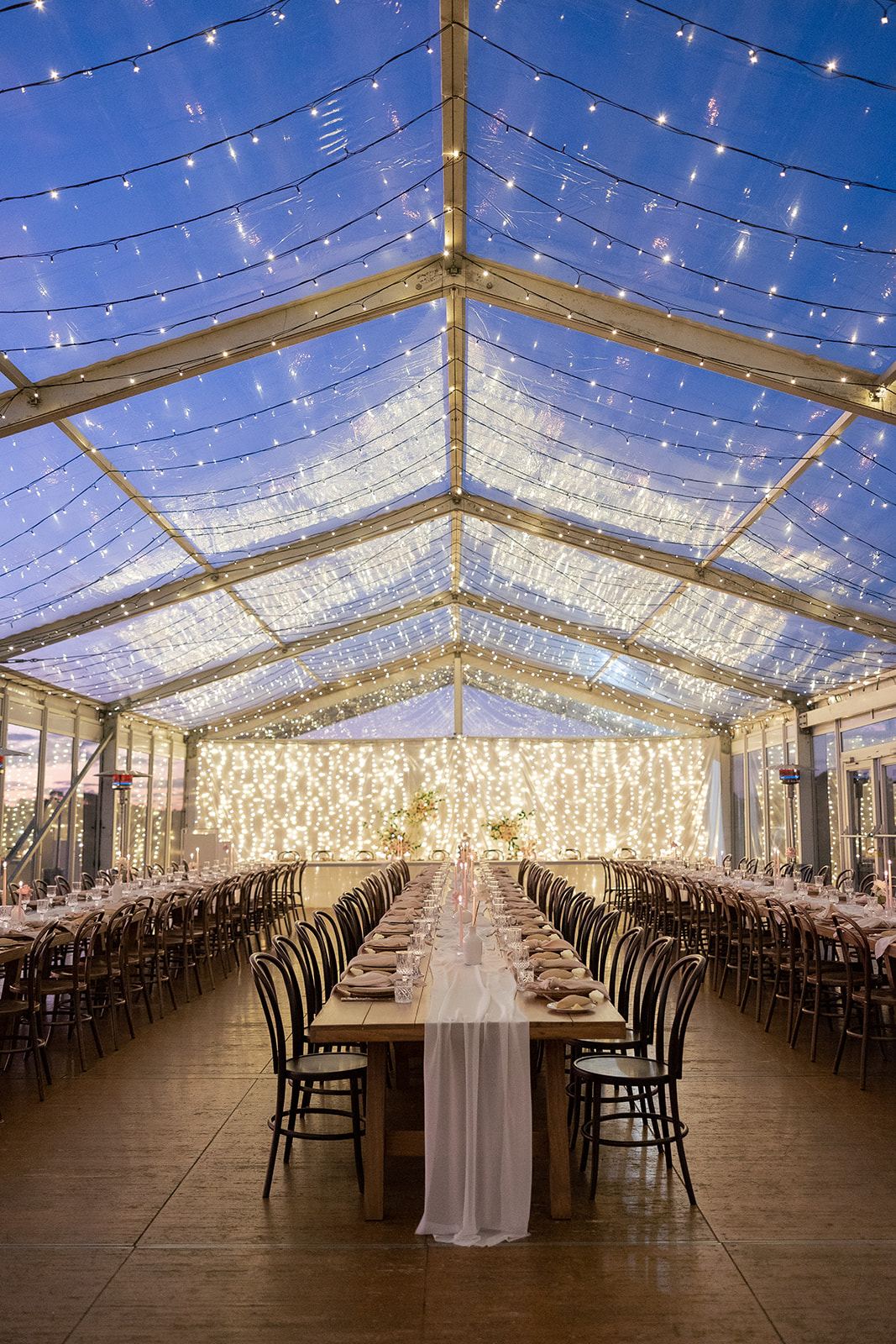 A long table under the clear marquee roof, strung with fairy lights at night