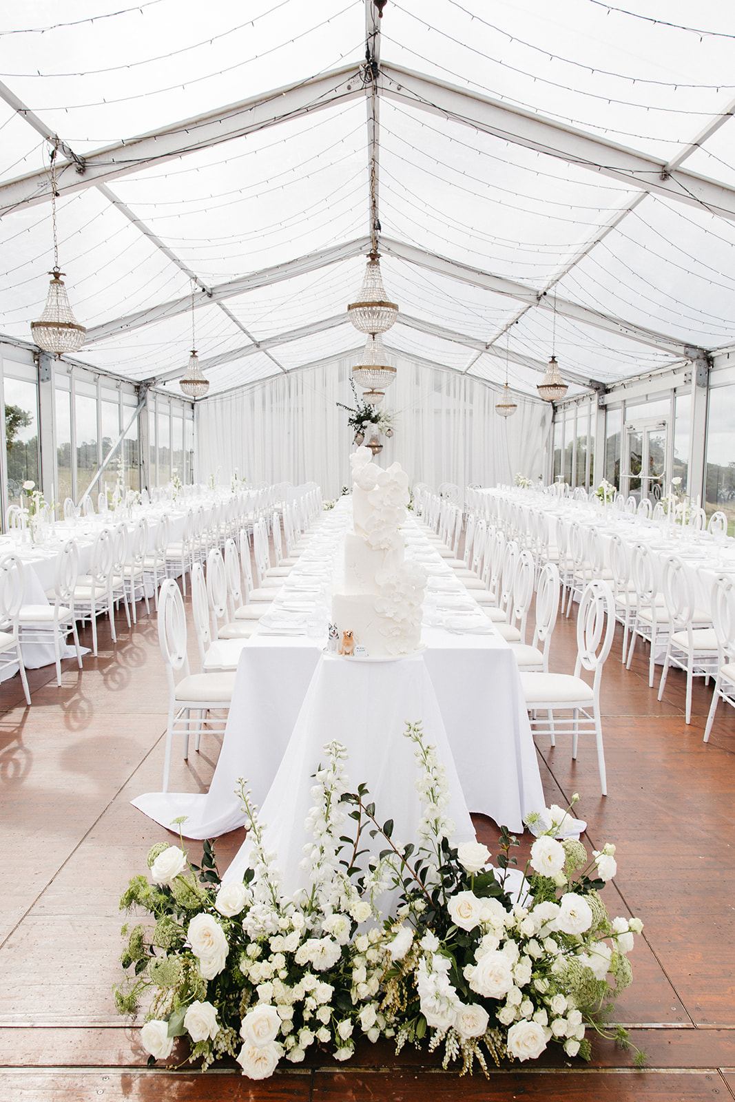 The marquee styled all in white, chandeliers overhead and the cake at the head of the long tables